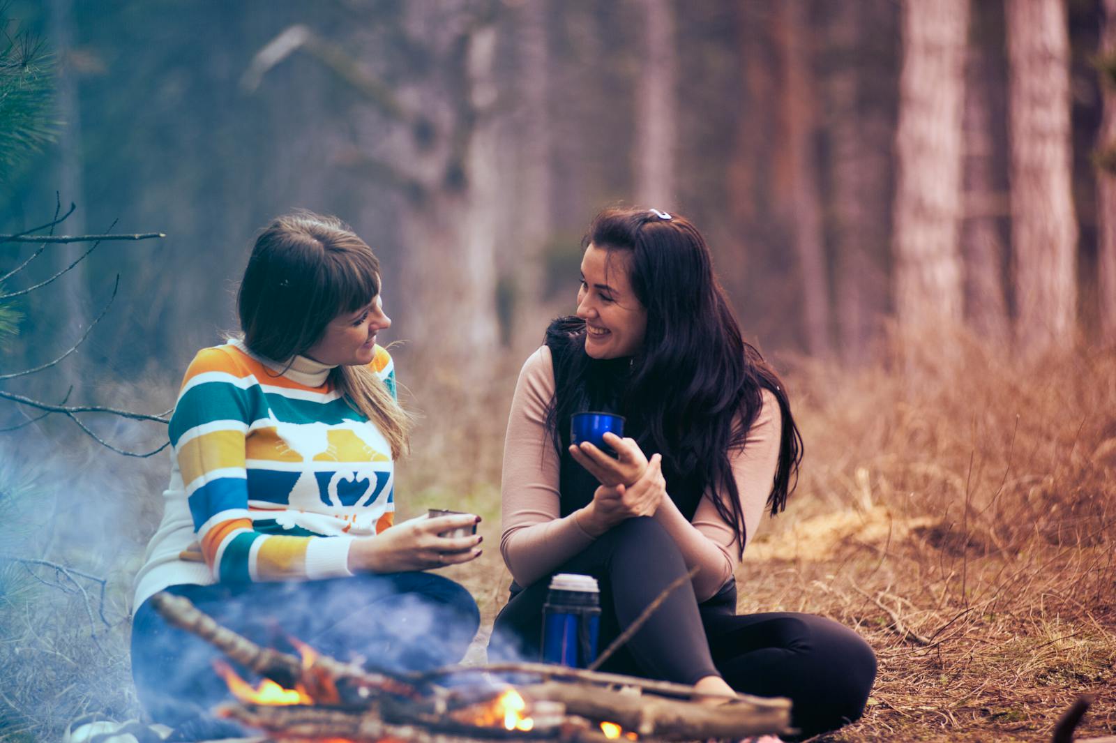 Two women enjoy coffee by a campfire in a forest, embracing the warmth and togetherness of nature.